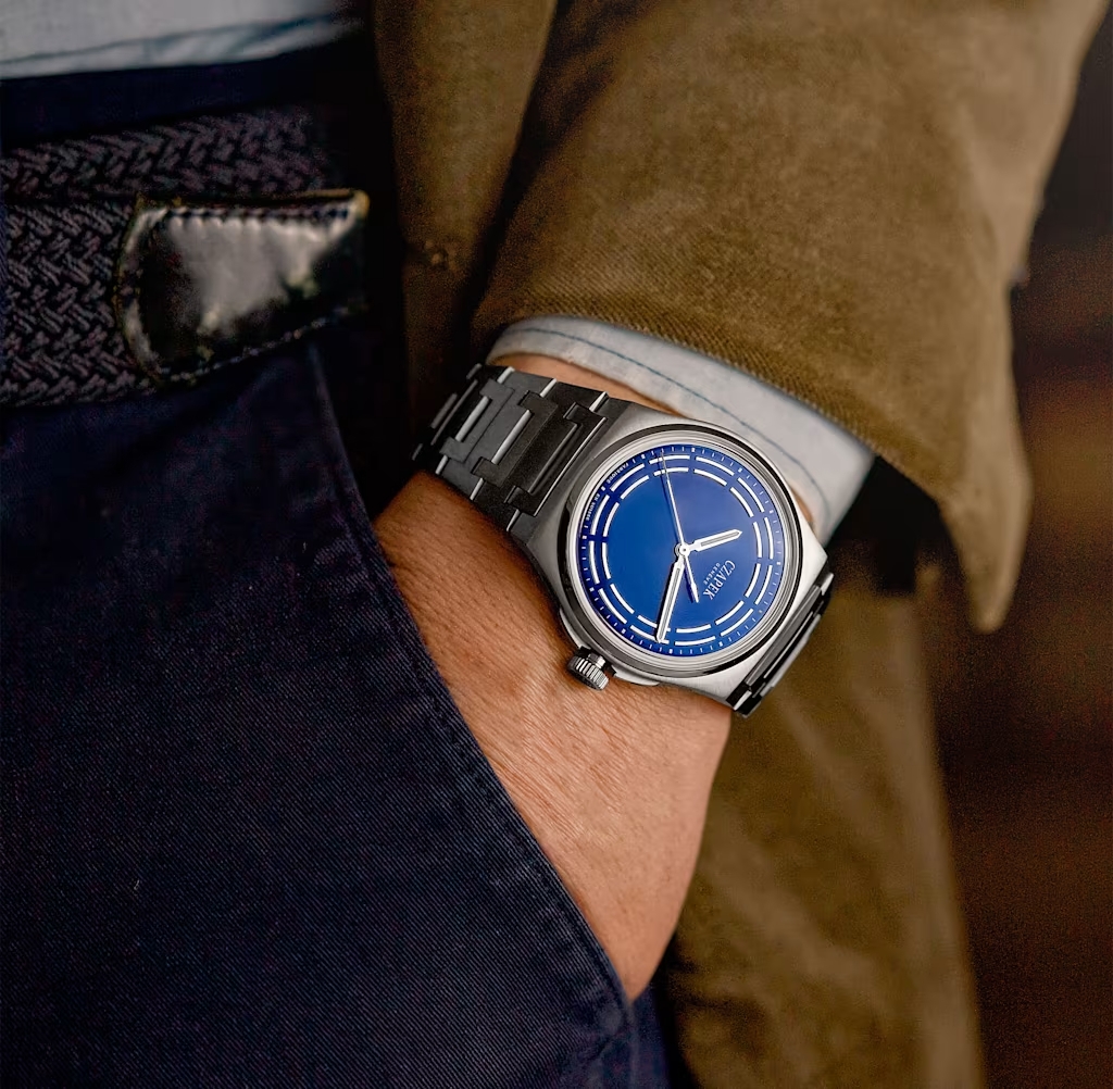Ein Mann trägt am Handgelenk eine silberne Armbanduhr mit blauem Zifferblatt und Metallarmband, dazu eine braune Jacke und dunkelblaue Hosen.