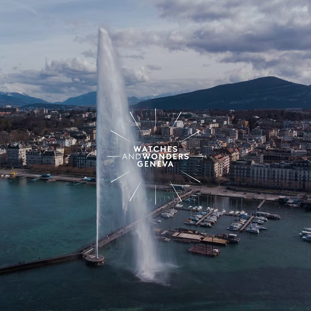 In einer Stadt mit Bergen im Hintergrund schießt eine hohe Wasserfontäne aus einem See hervor, darüber ist der Text „WATCHES AND WONDERS GENEVA“ eingeblendet.