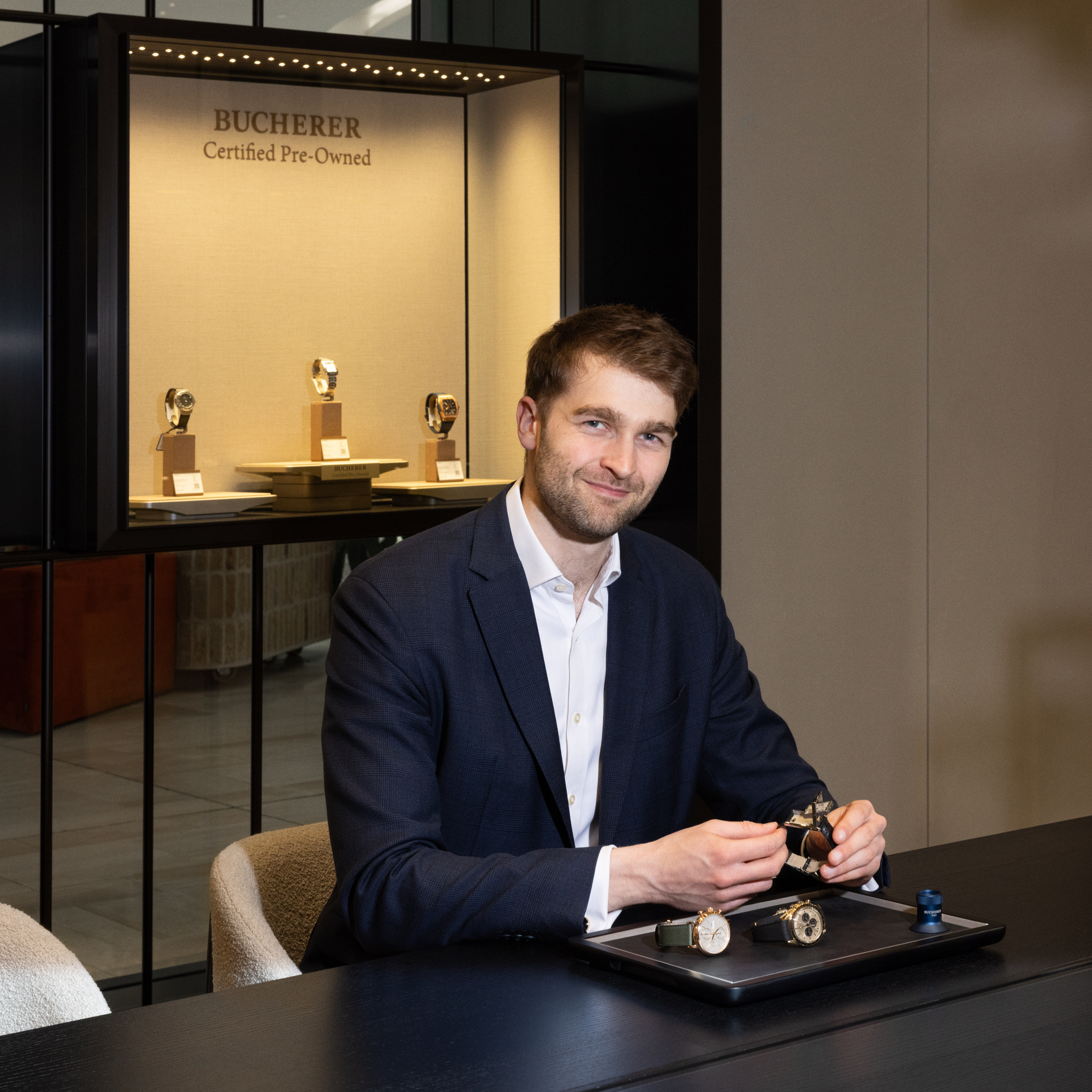 A man in a suit sits at a table with watches, smiling at the camera, with a Bucherer display case behind him.