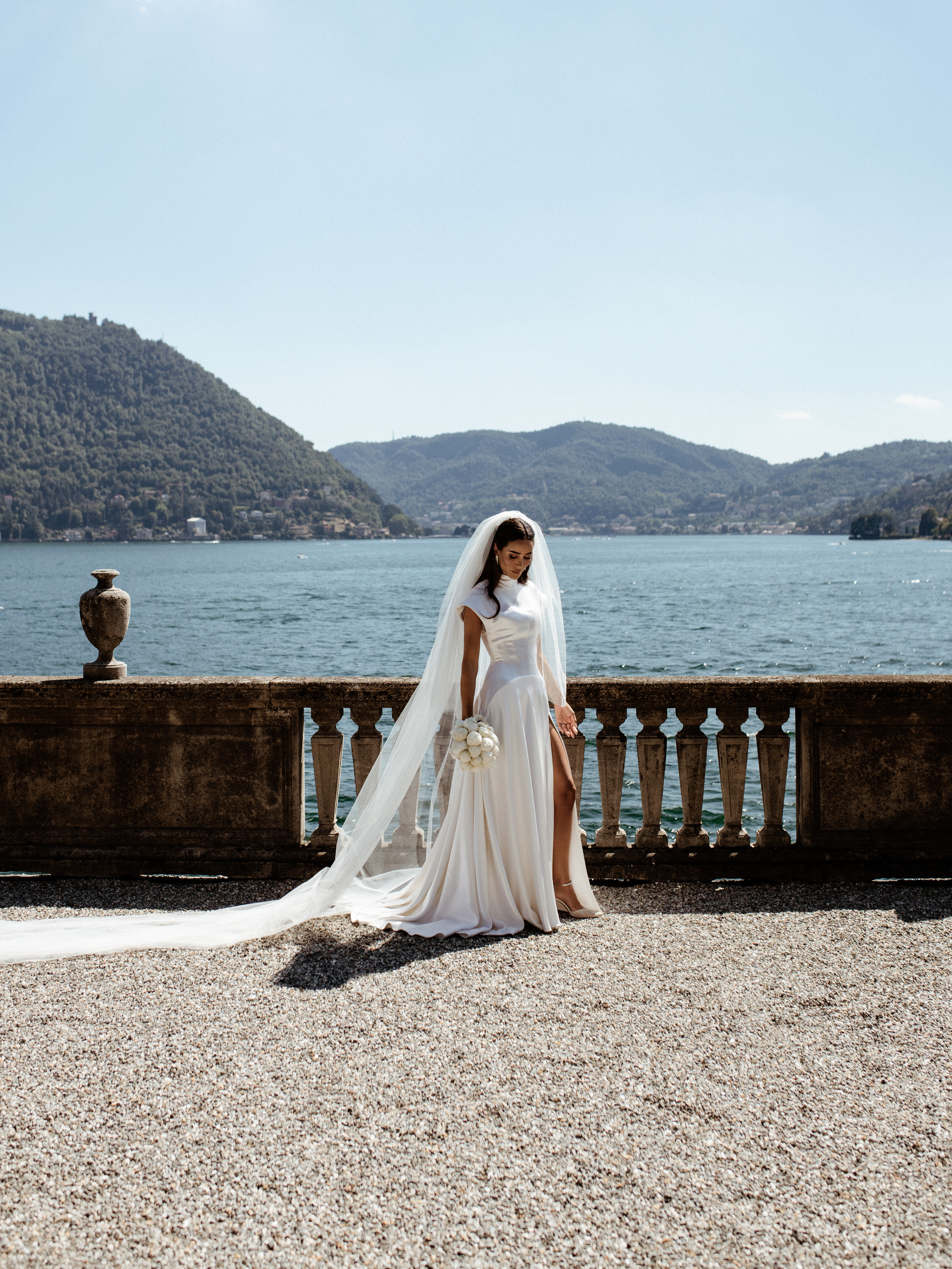 Eine Braut in einem weißen Kleid und langem Schleier steht auf einer Steinterrasse mit Blick auf einen See und die Berge.