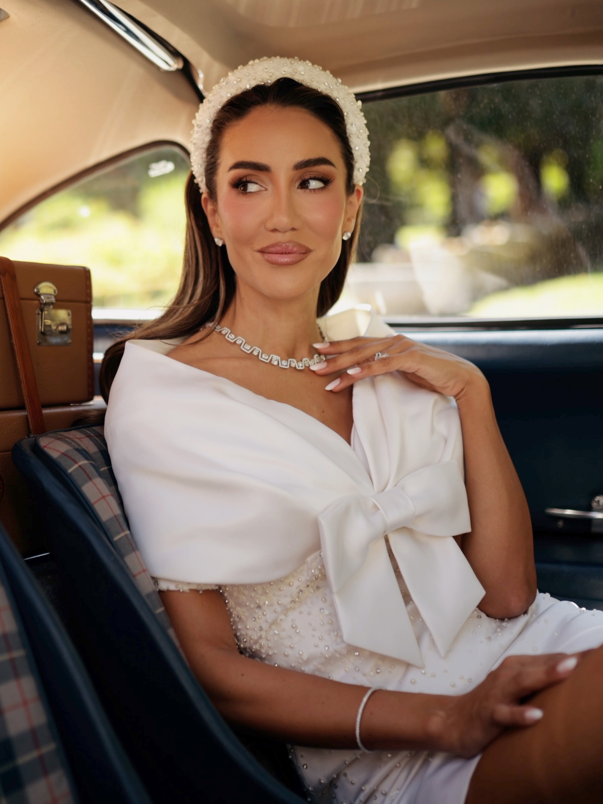 Femme en robe blanche et bandeau orné de perles, portant un collier de diamants, assise dans une voiture.