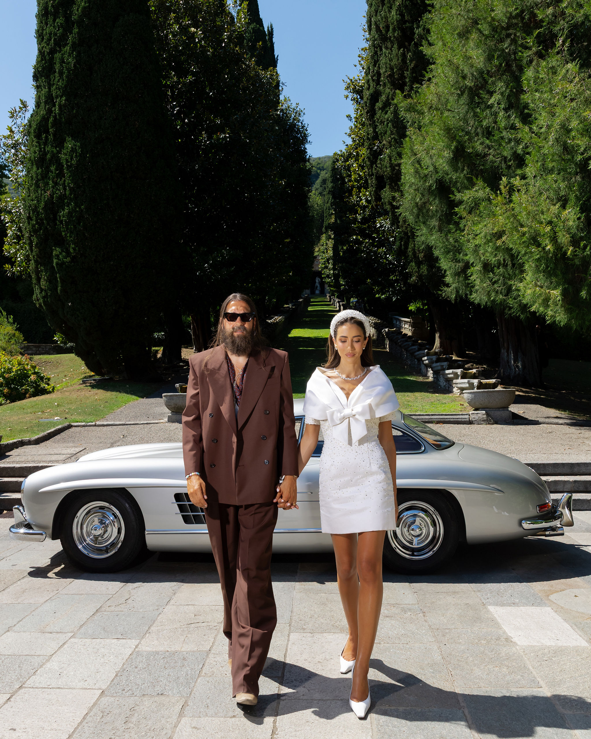 Ein Mann in einem braunen Anzug und eine Frau in einem weißen Kleid gehen Hand in Hand vor einem silbernen Oldtimer entlang.