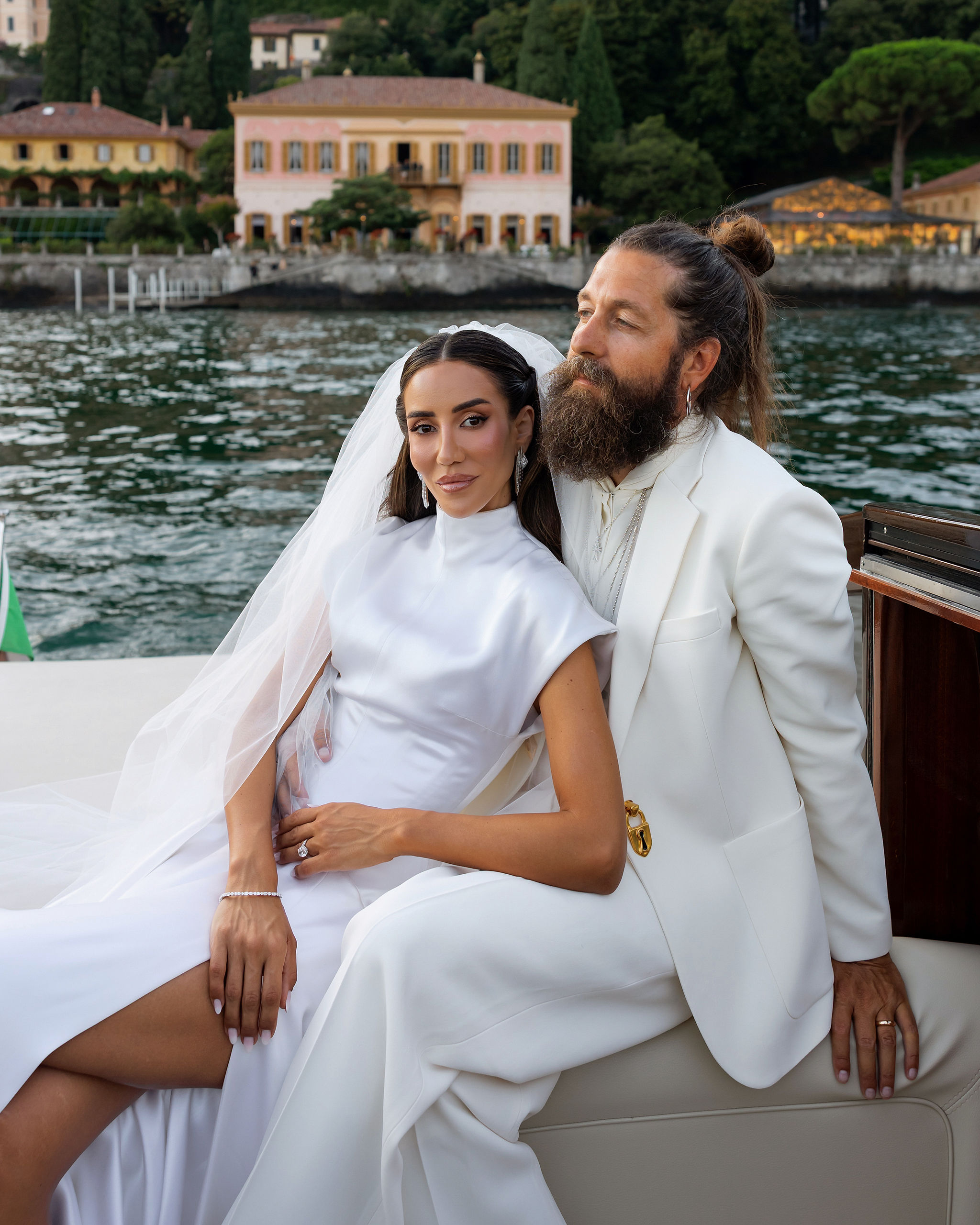 Eine Braut in einem weißen Kleid und ein Bräutigam in einem weißen Anzug sitzen auf einem Boot; im Hintergrund ist eine große Villa zu sehen.