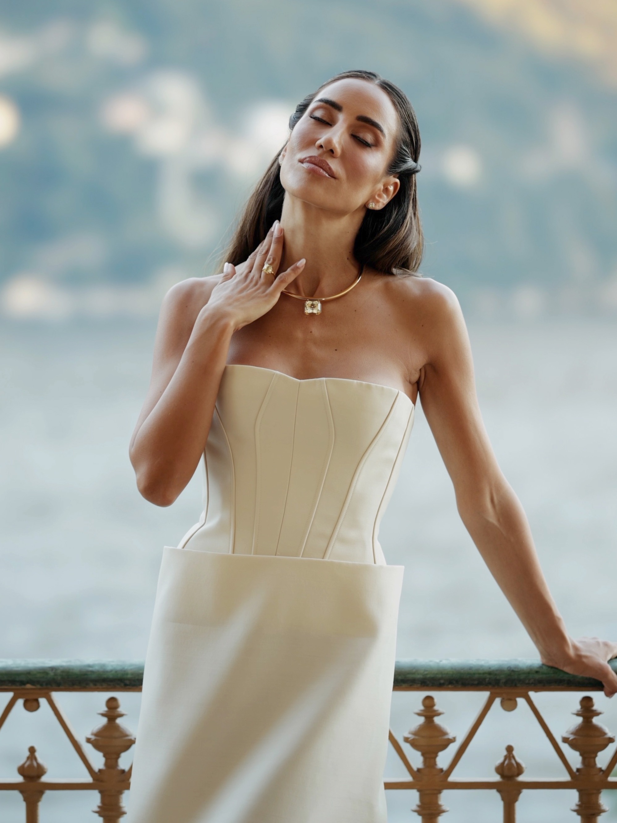 Eine Frau in einem trägerlosen cremefarbenen Kleid und einer goldenen Halskette mit einem großen quadratischen Anhänger steht auf einem Balkon mit Blick aufs Wasser.