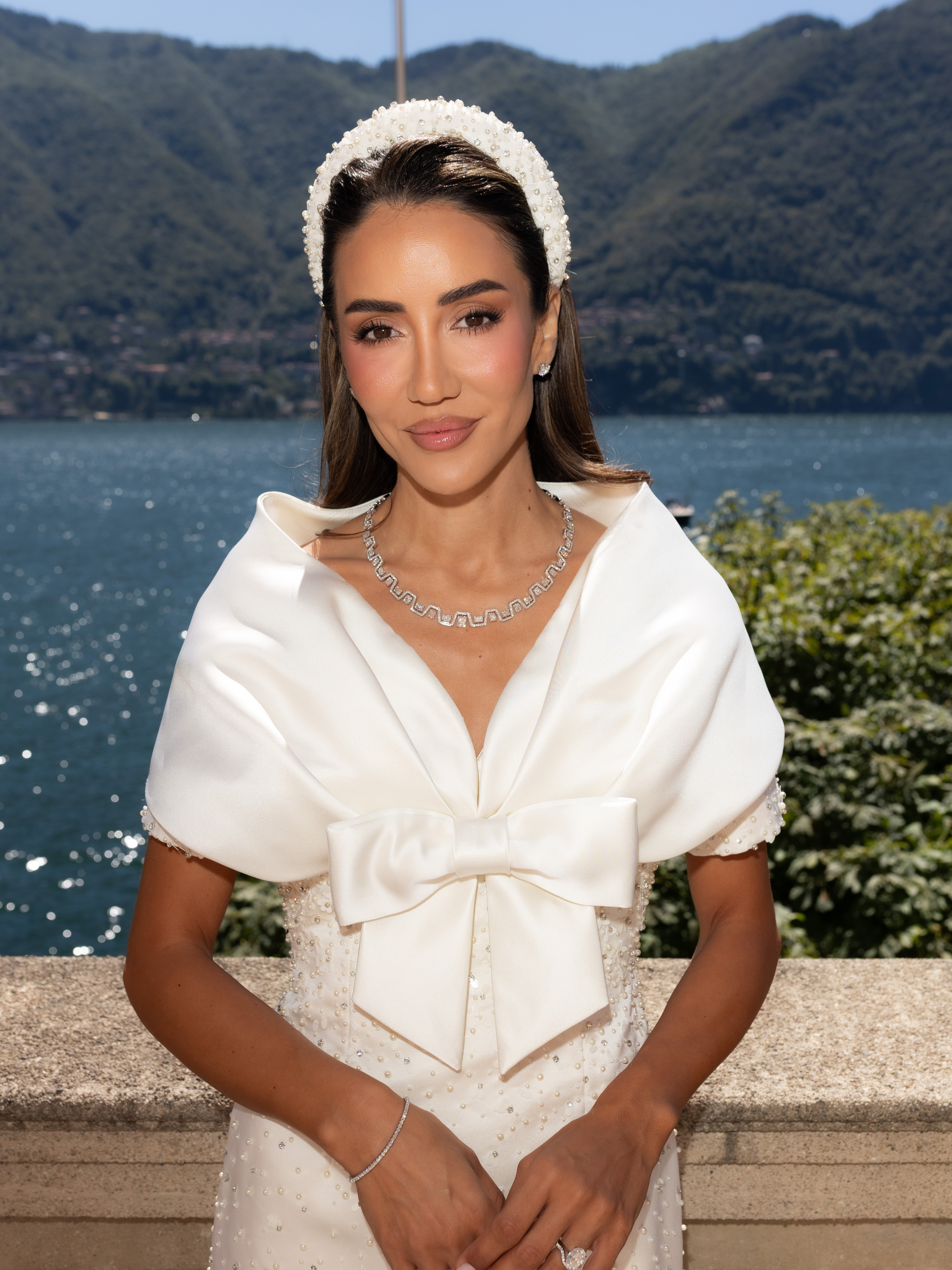 Eine Frau in einem weißen Kleid mit einer großen Schleife und einem Perlenstirnband steht im Freien vor einem See und Bergen im Hintergrund.