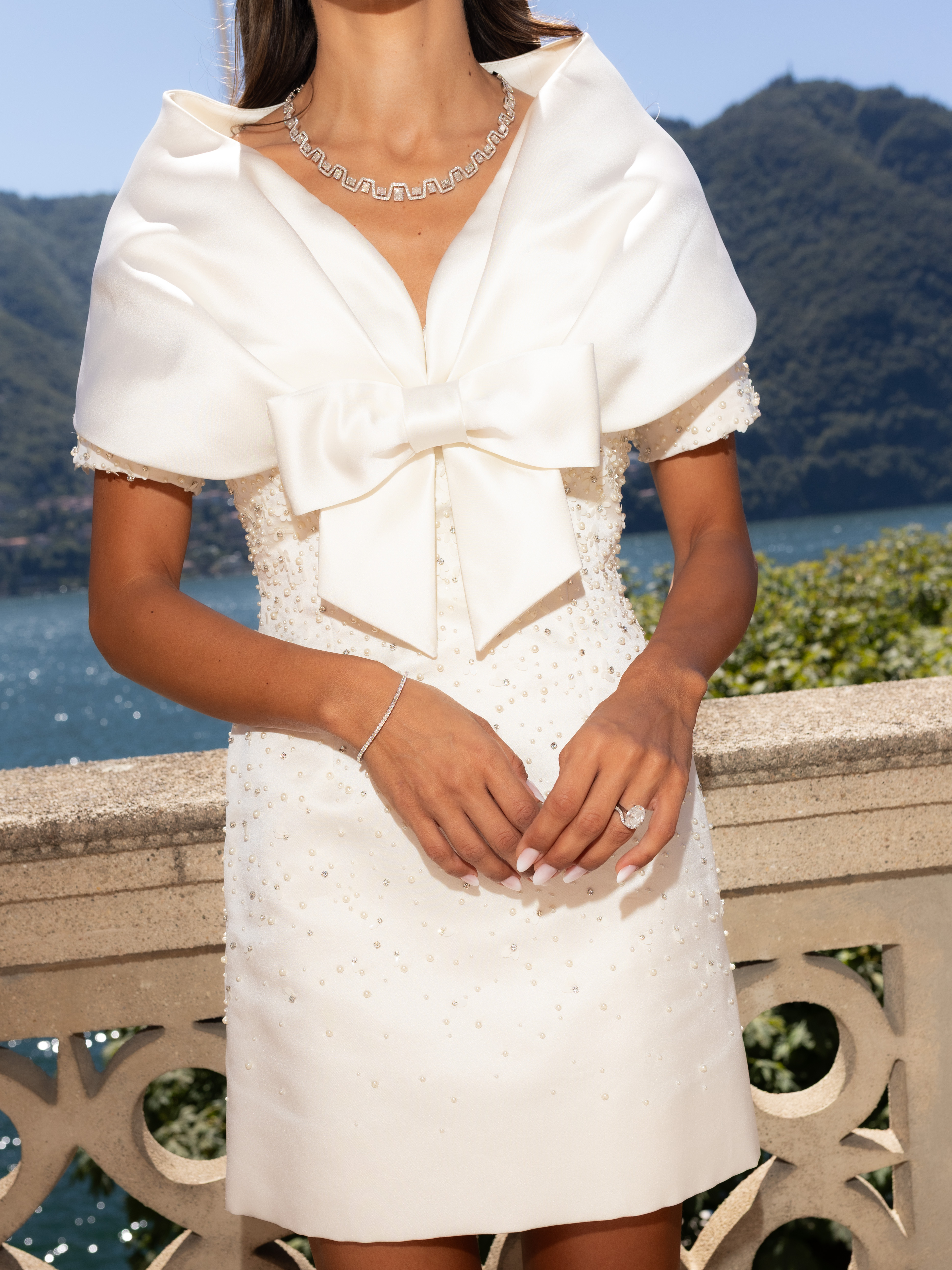 Eine Frau in einem weißen, kurzen, schulterfreien Kleid mit großer Schleife und Perlenverzierungen, die eine Diamantkette, ein Armband und einen Ring trägt, steht auf einem Balkon mit Blick auf einen See und Berge.