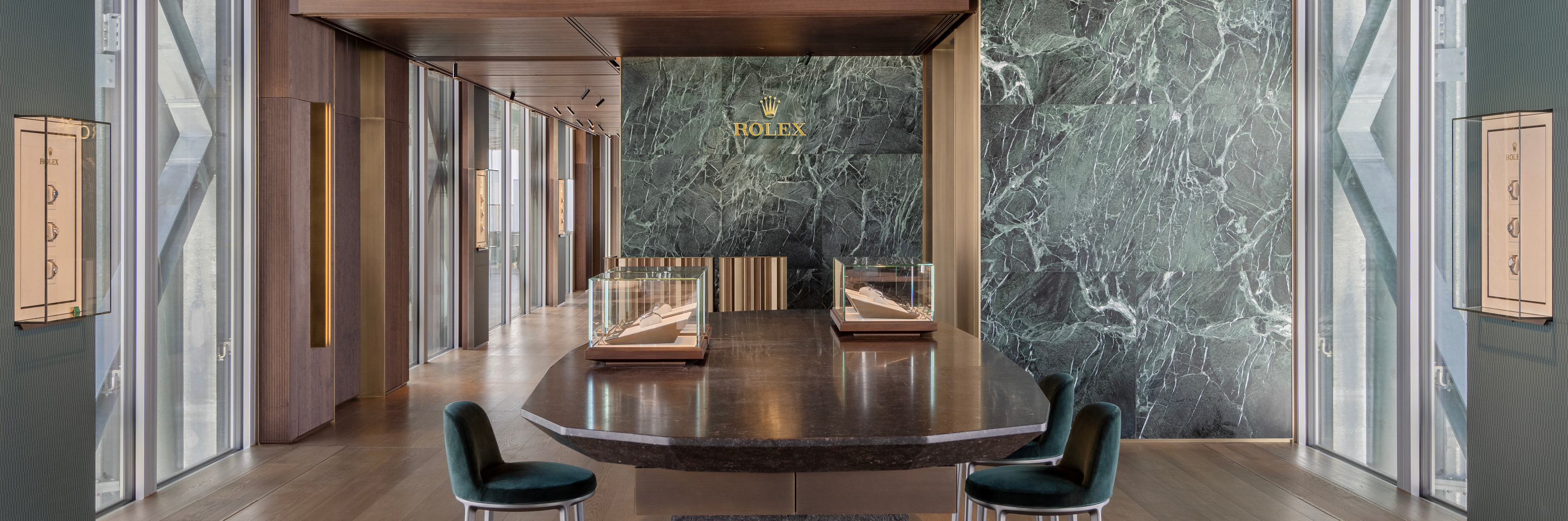Das luxuriöse Interieur eines Rolex-Geschäfts mit einem dunklen Marmortisch, zwei Vitrinen und einer grünen Marmorwand mit dem Rolex-Logo.