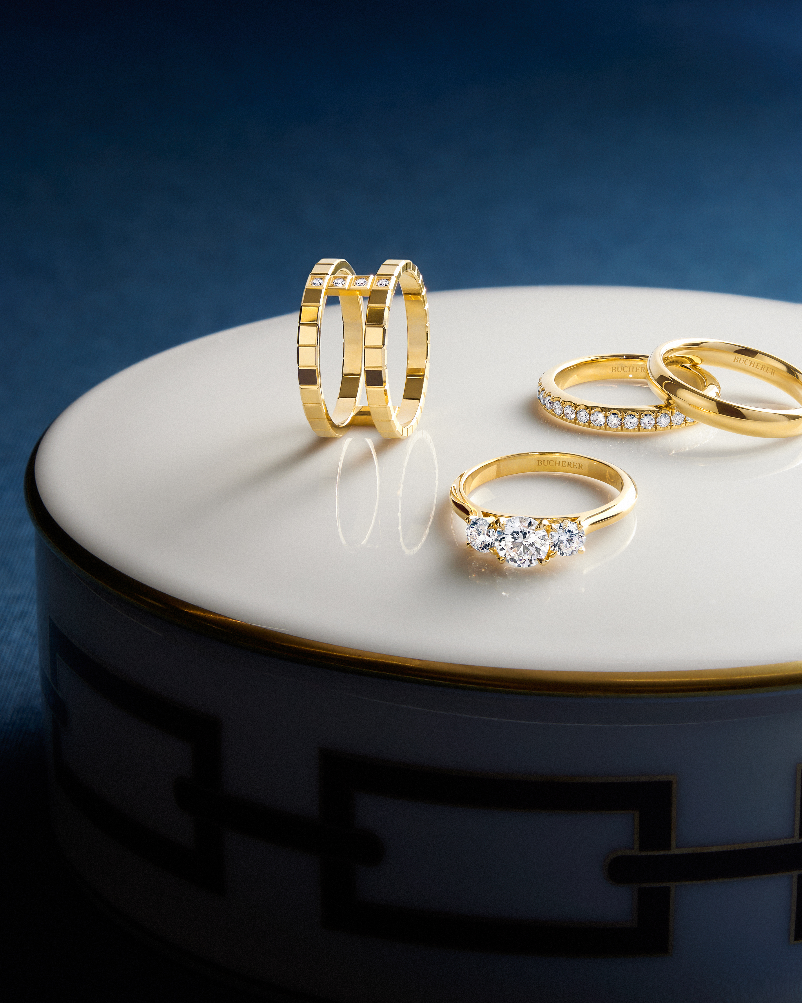 Four gold rings, including diamond bands and a three-stone ring, displayed on a white and gold surface.