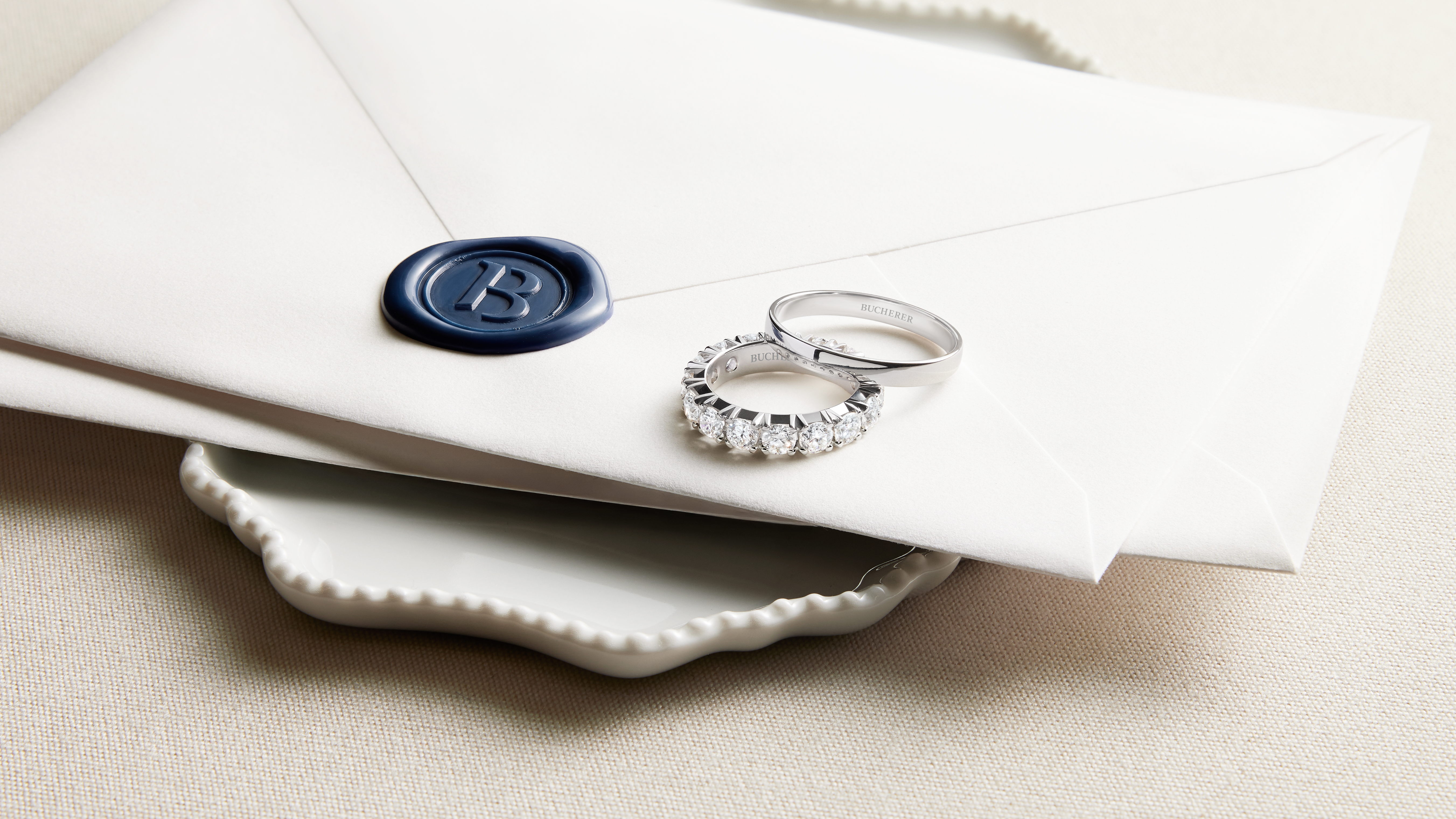 Two diamond rings rest on a white envelope sealed with a blue wax stamp featuring the letter B.