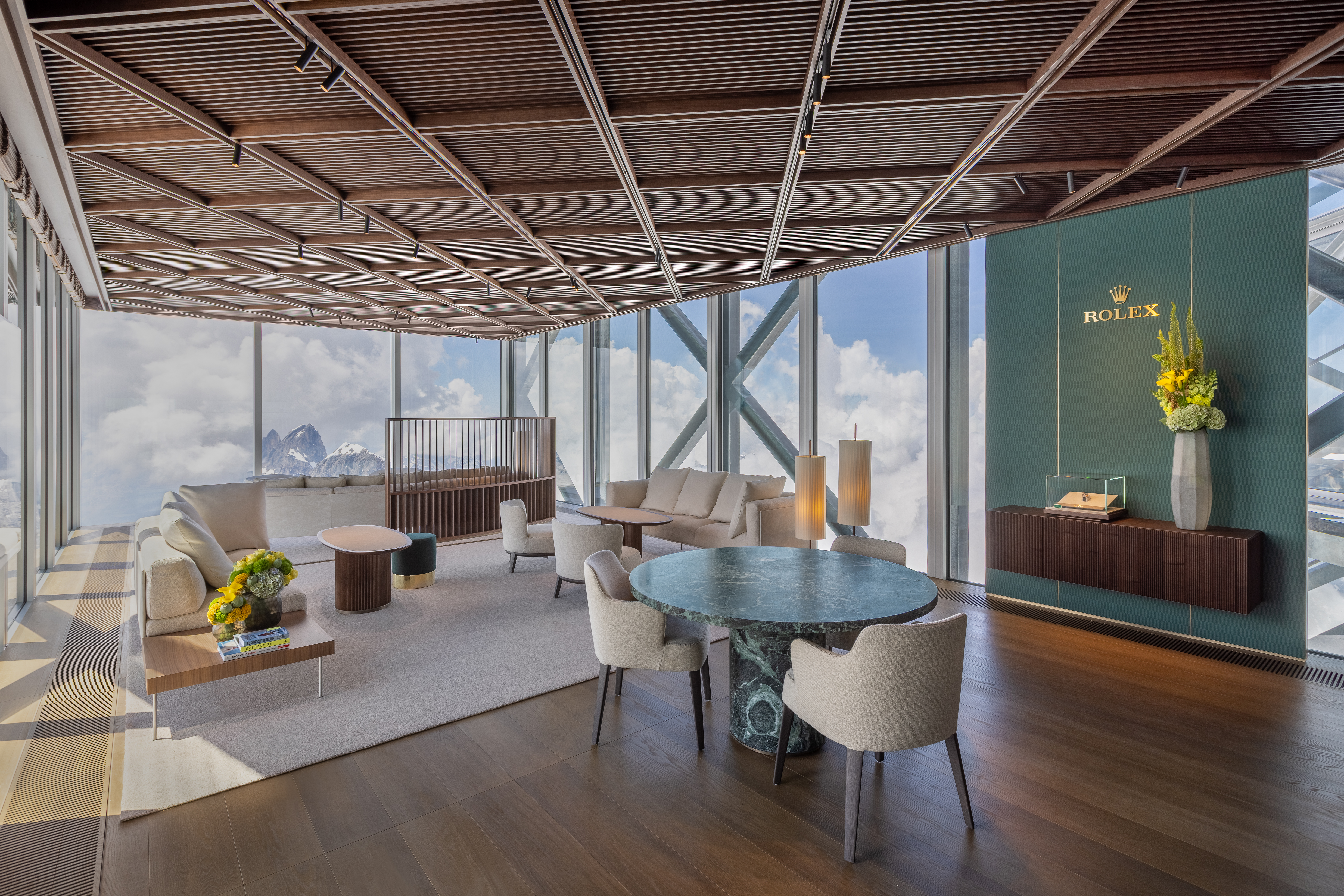 Un salon moderne avec de grandes fenêtres donnant sur les montagnes, comprenant des canapés, des fauteuils, des tables et une vitrine Rolex.