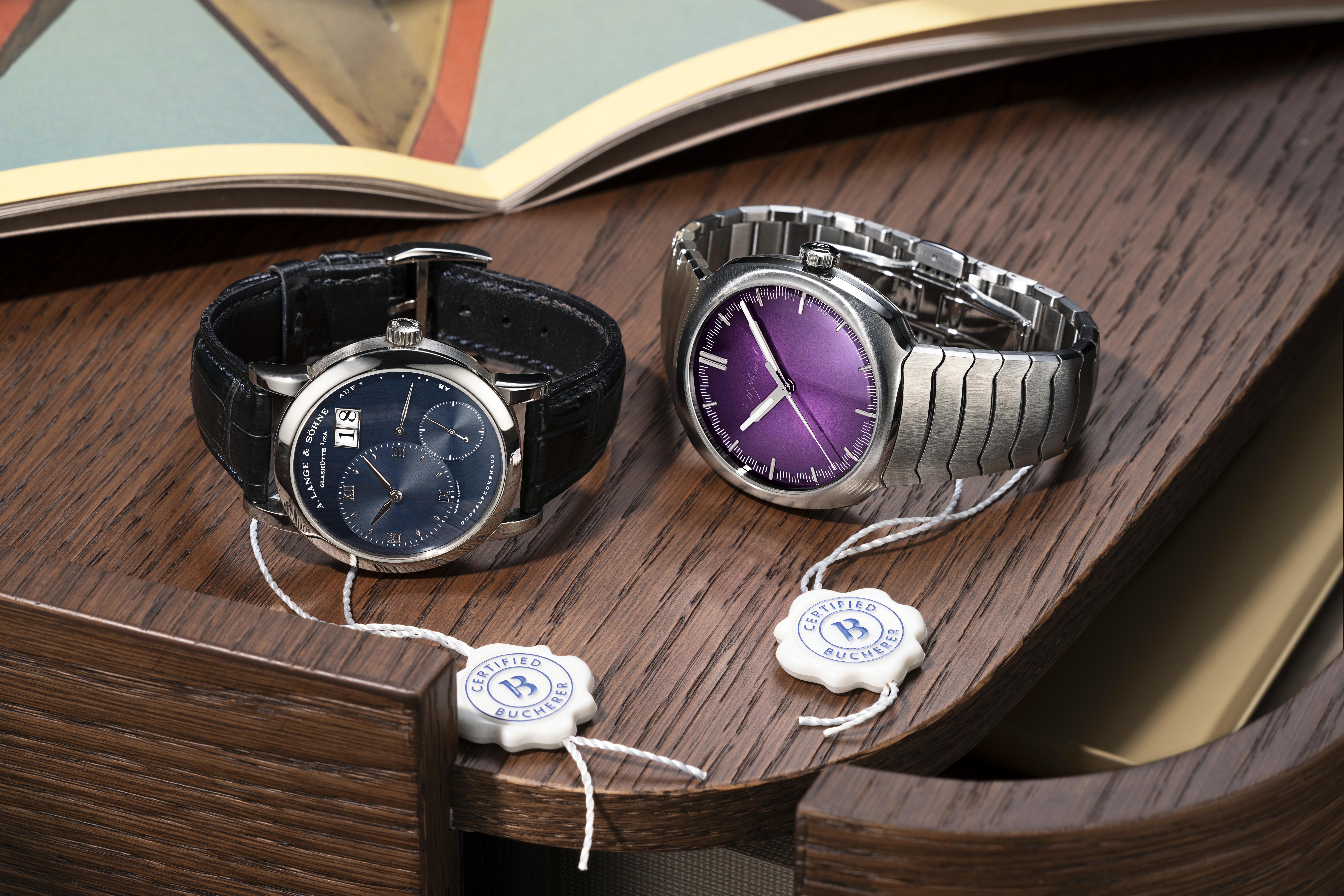 Zwei Luxusuhren, eine mit blauem Zifferblatt und Lederarmband, die andere mit violettem Zifferblatt und Metallarmband, auf einer Holzoberfläche.