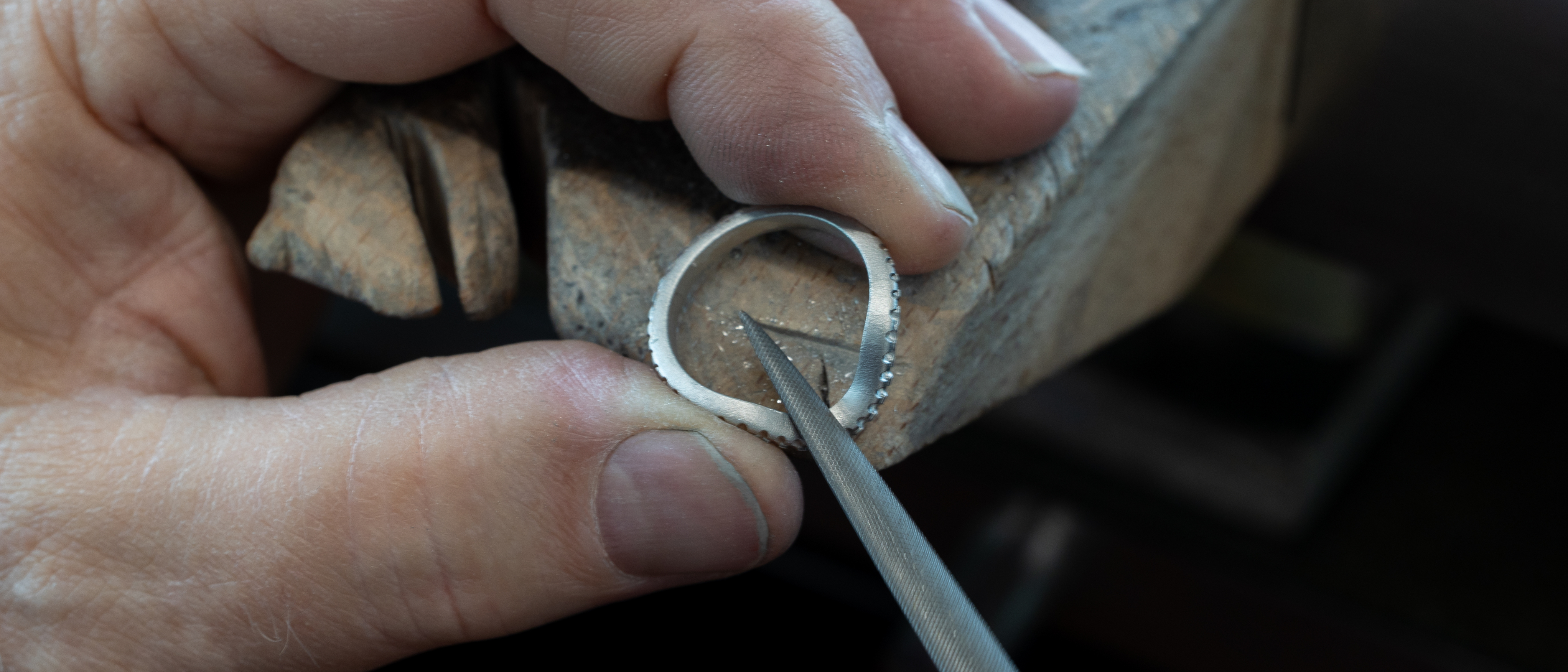 A jeweler's hands filing the inside of a silver ring with small diamonds on a wooden workbench.