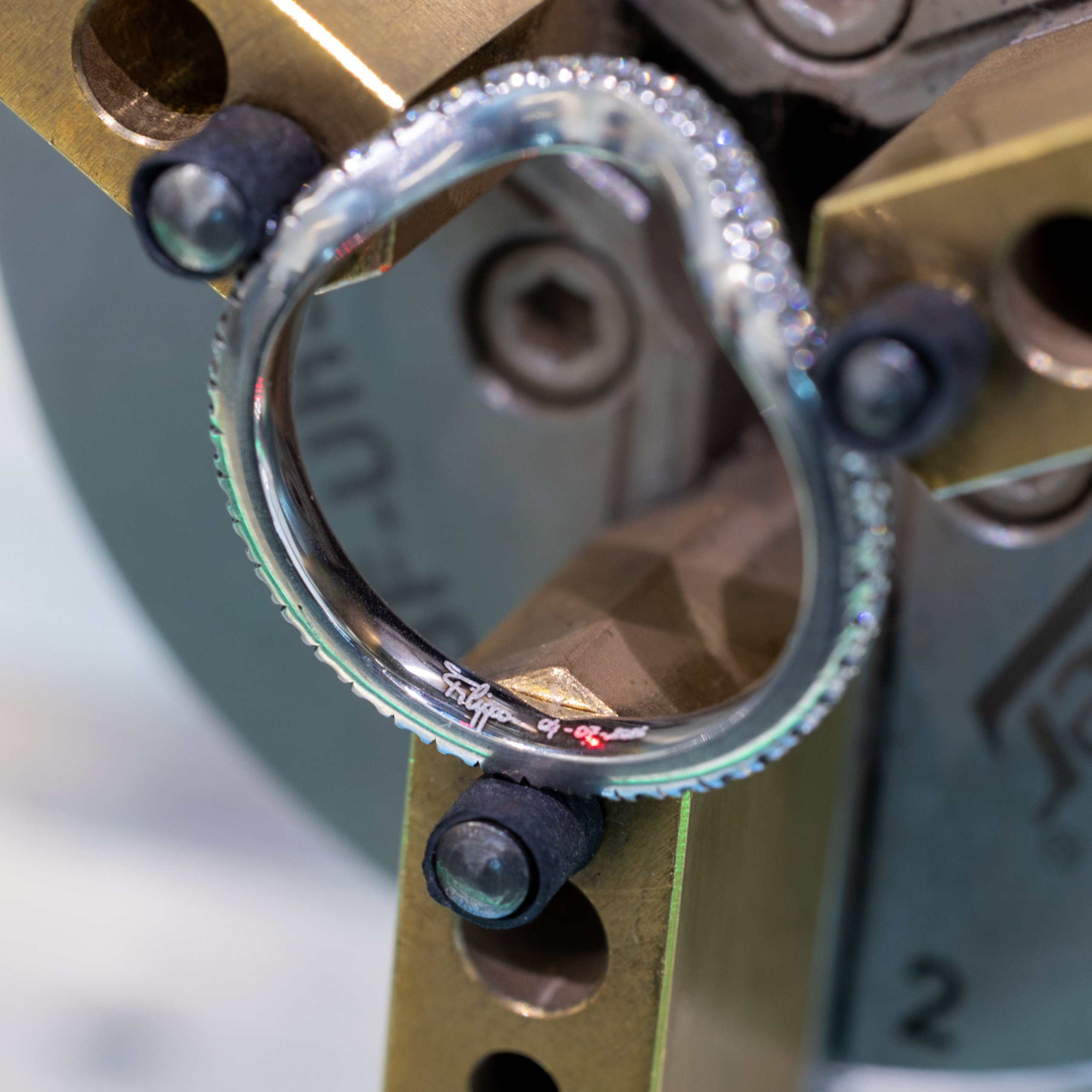 A diamond-encrusted ring with an engraved inscription, held by a metal clamp.