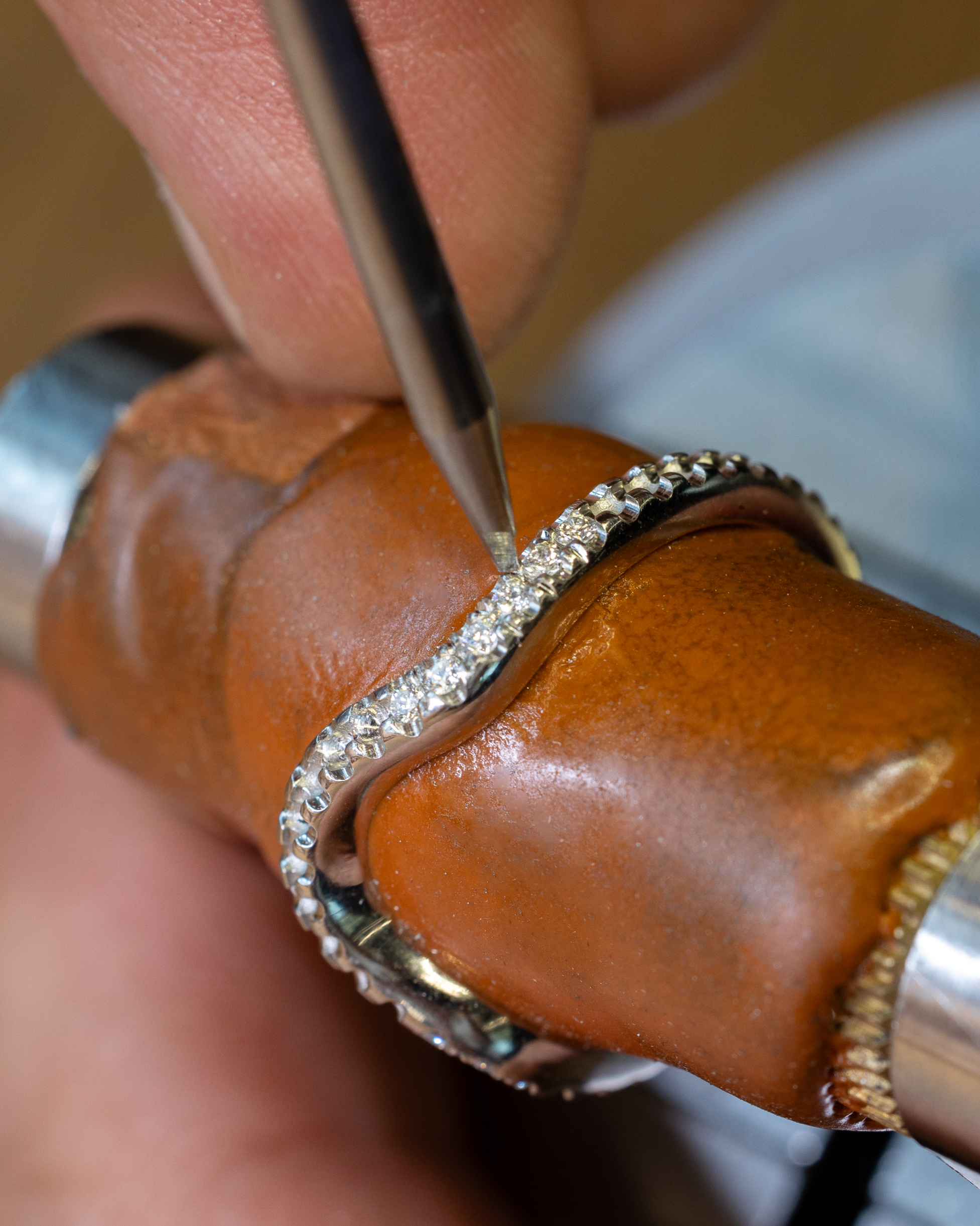 A jeweler's hand uses a tool to set small diamonds into a silver ring on a brown ring mandrel.