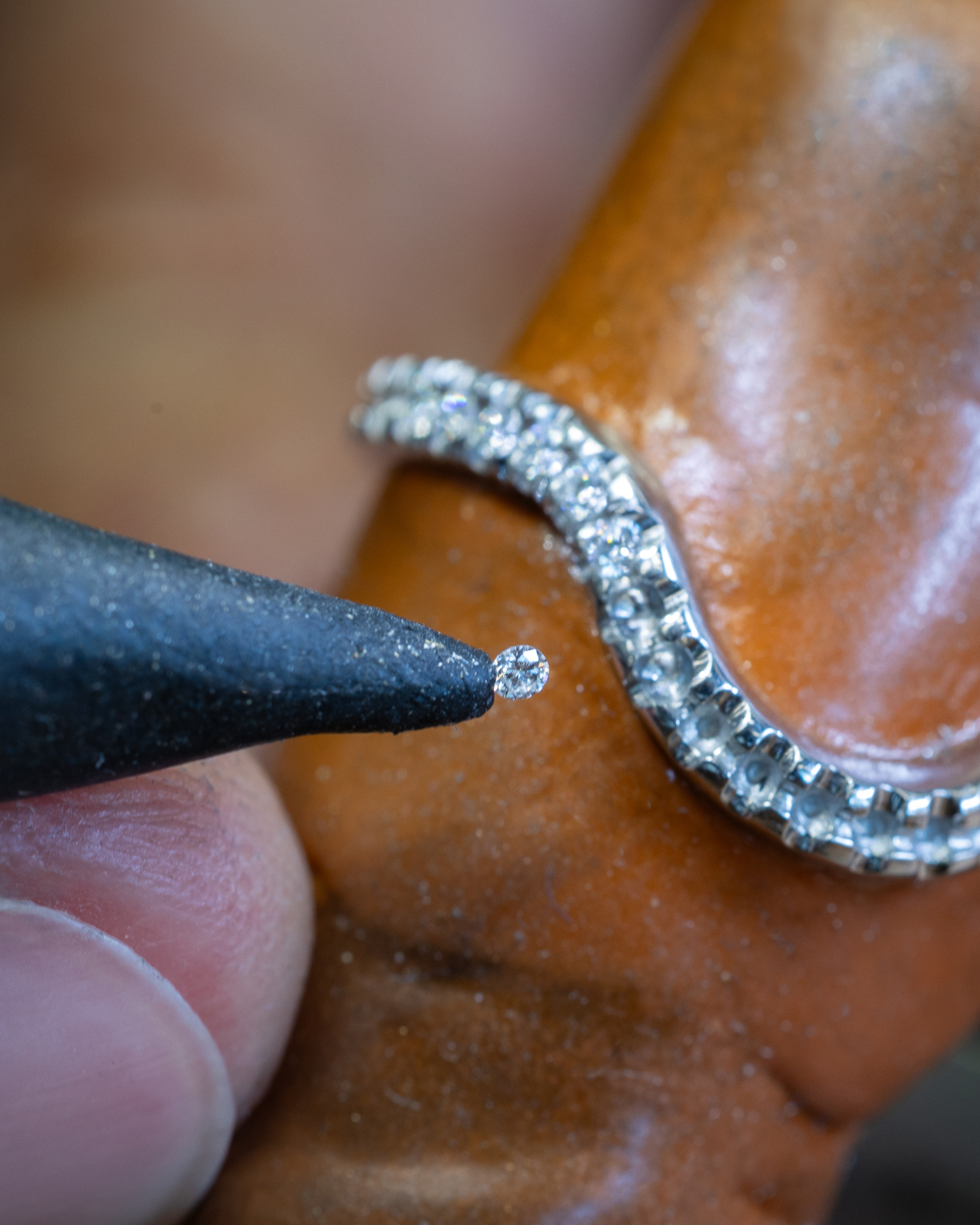 A jeweler's hand holds a tiny diamond with tweezers, preparing to set it into a diamond-encrusted ring on a brown ring mandrel.