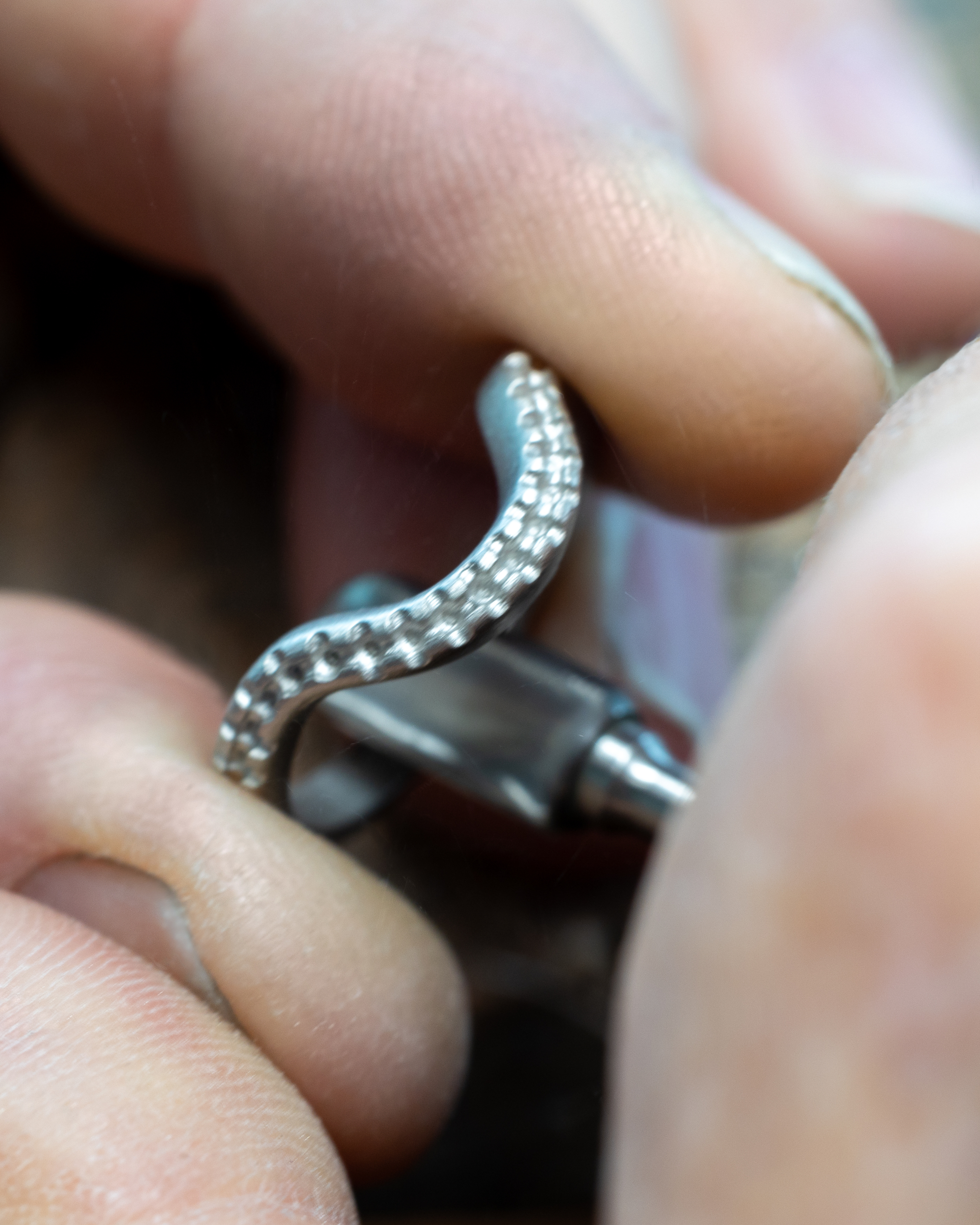 A jeweler's hands hold a silver ring with a textured, wavy design, working on it with a tool.