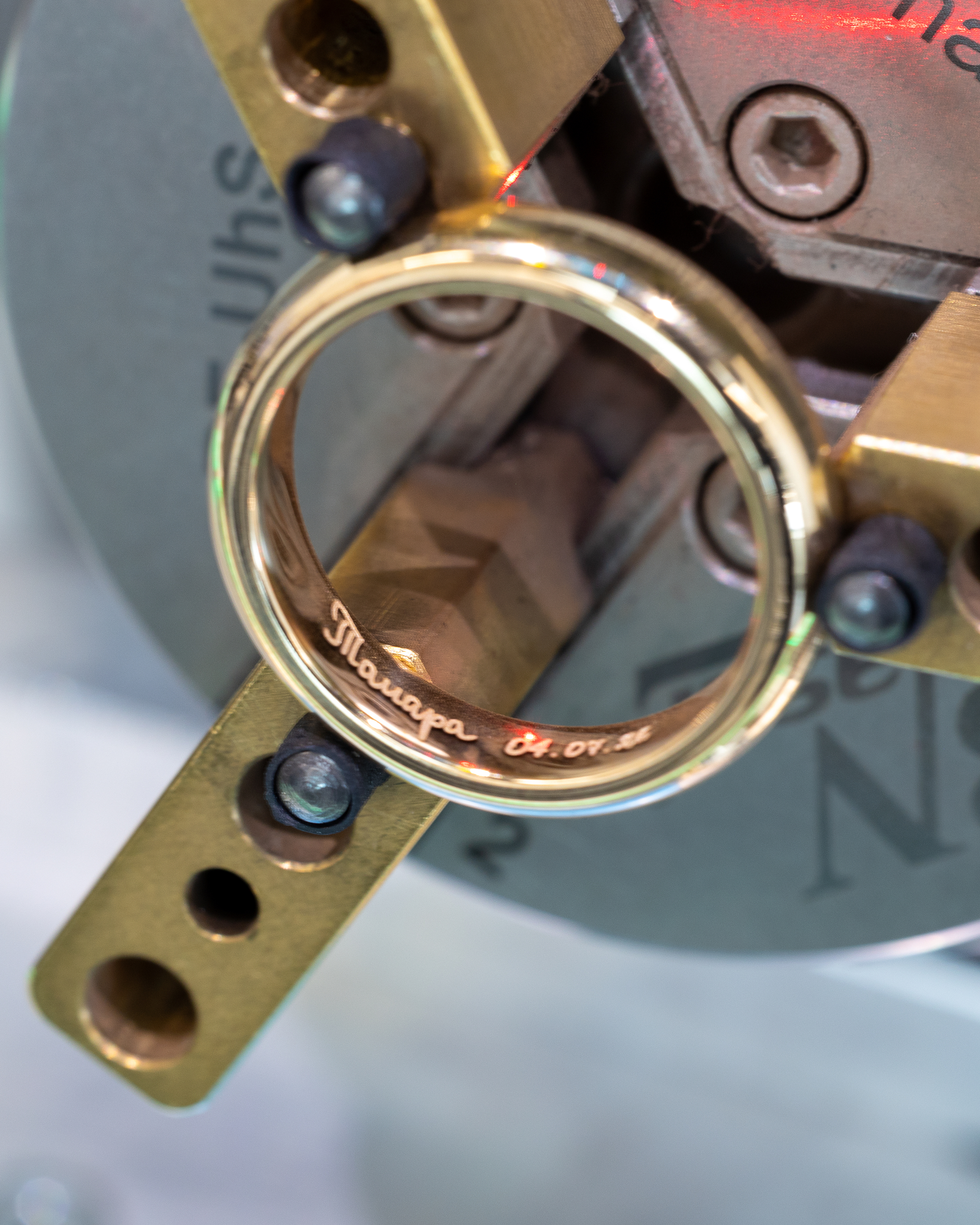 A gold ring with engraved text and numbers is held by a brass clamp, with a red laser light visible.