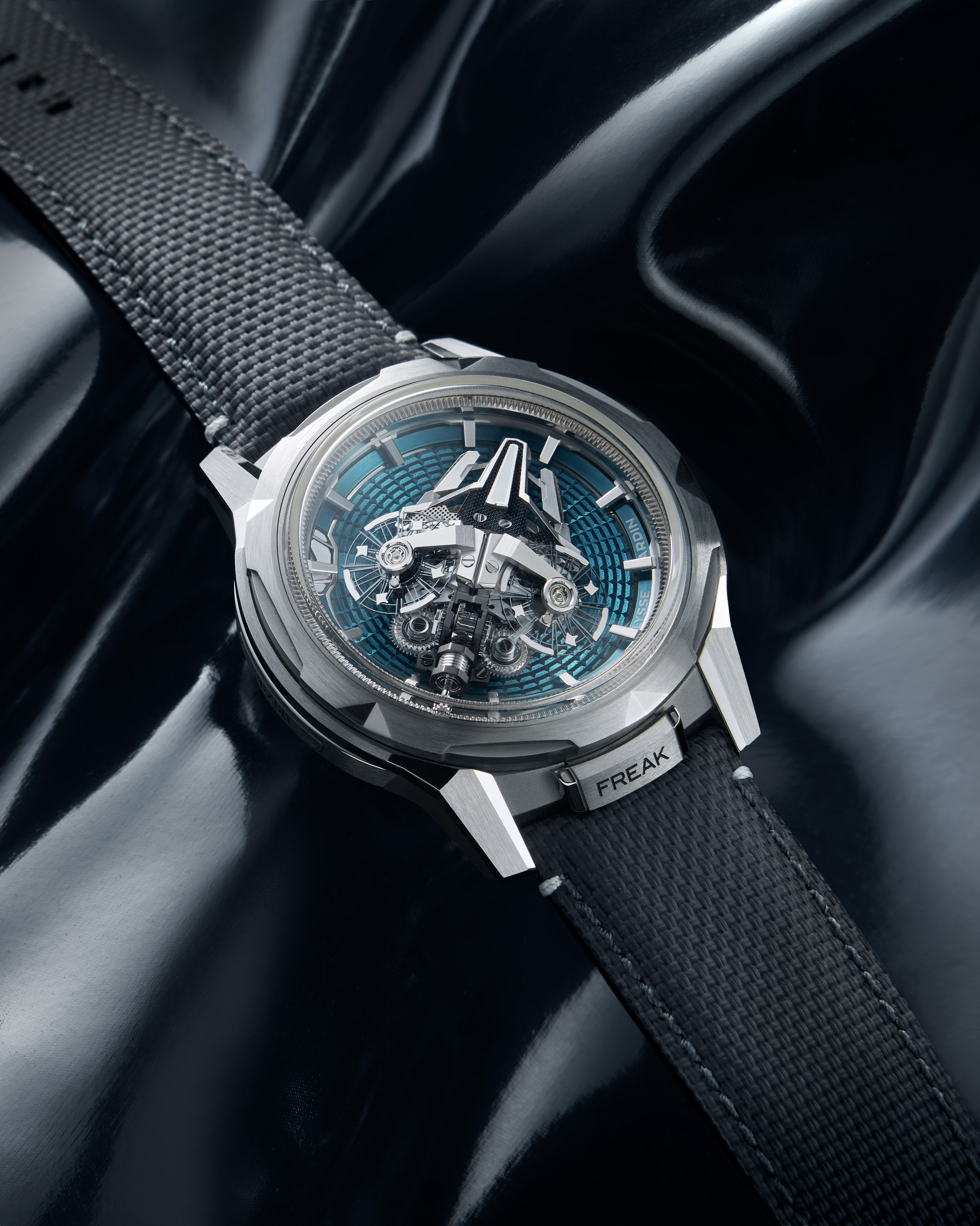 A silver watch with a blue and silver intricate face and a black textured band rests on a shiny black surface.