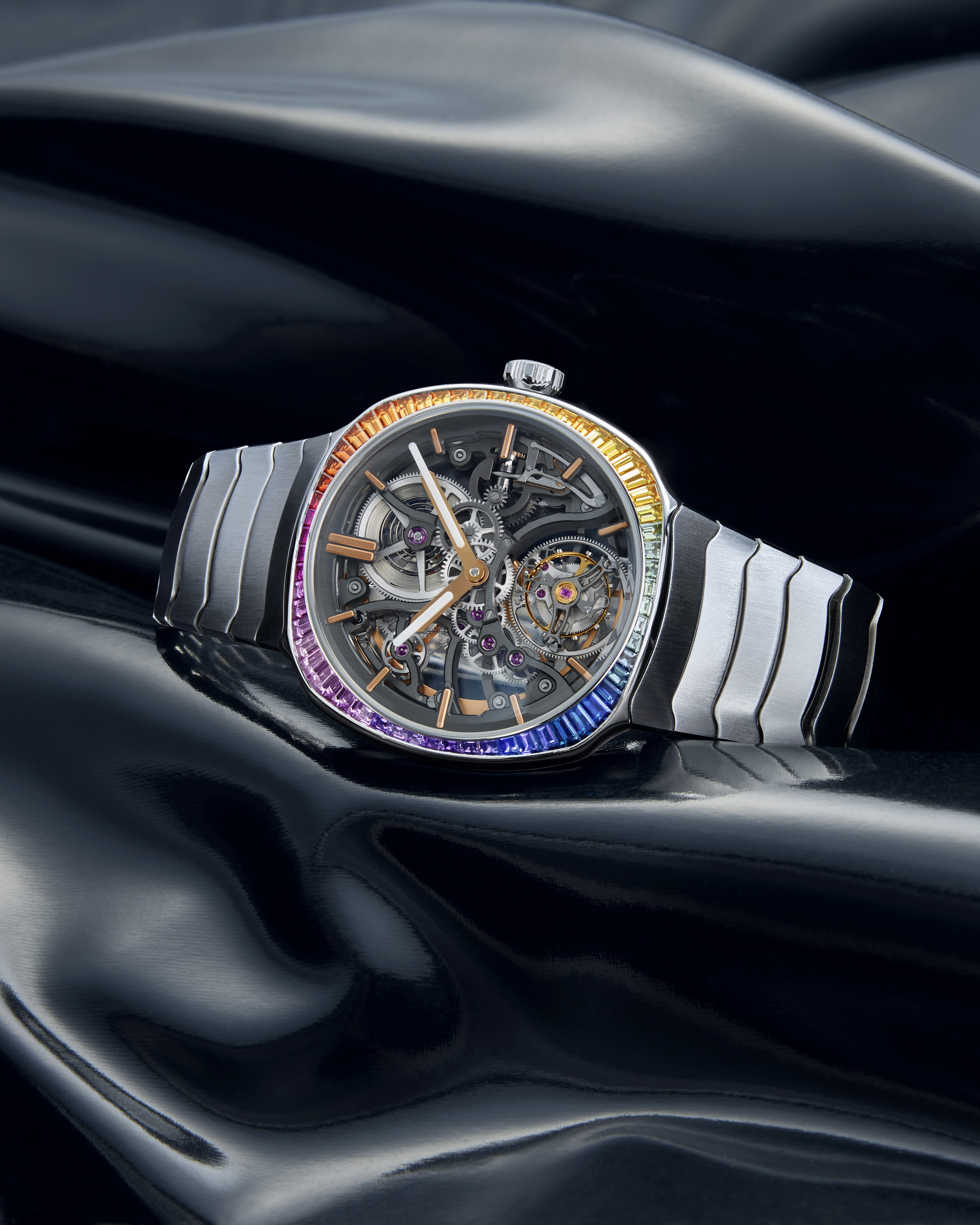 A luxury watch with a rainbow-gemmed bezel, exposed gears, and a metal bracelet, resting on a black satin surface.