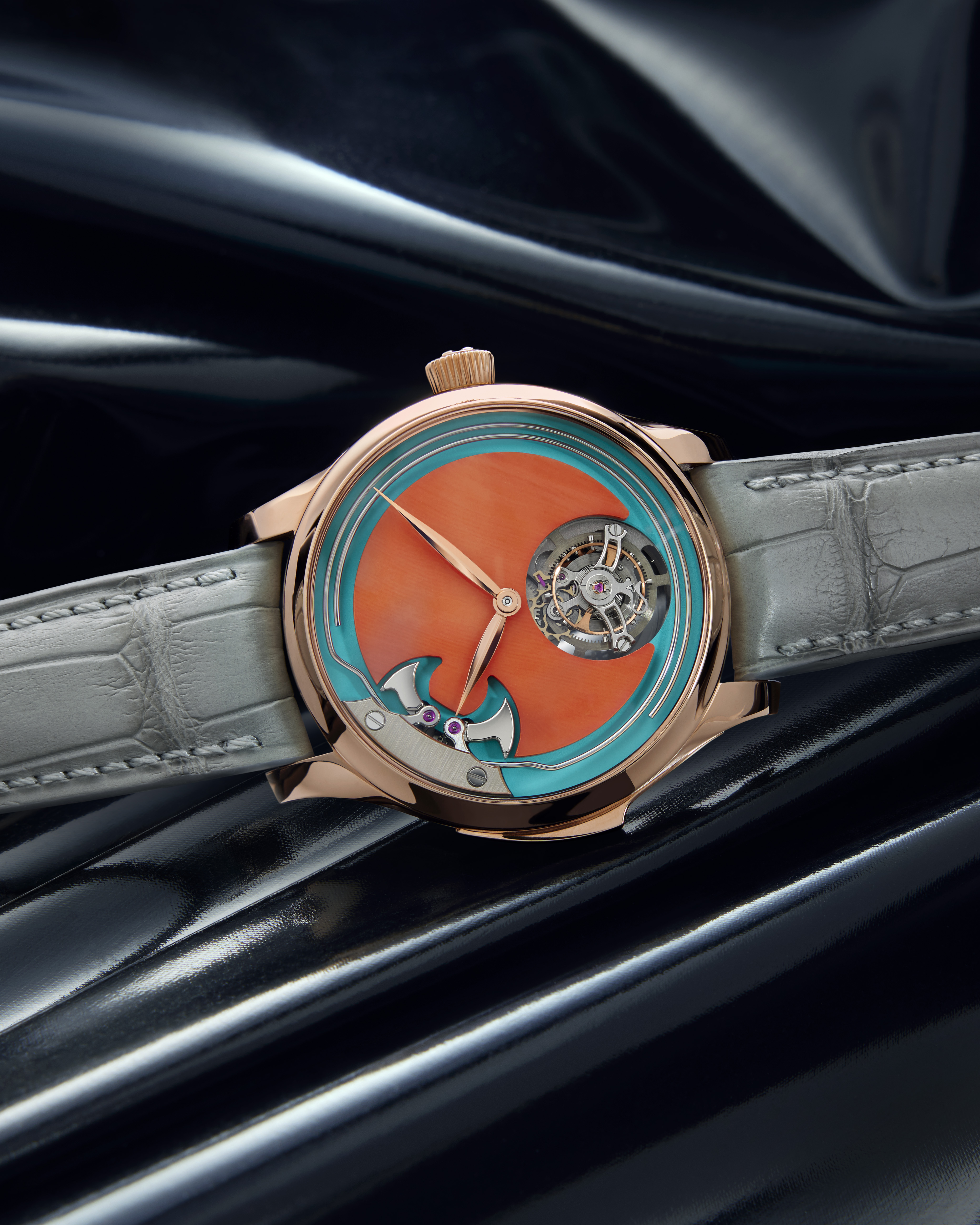 Rose gold watch with an orange and teal face, exposed gears, and a gray leather strap on a dark, reflective surface.