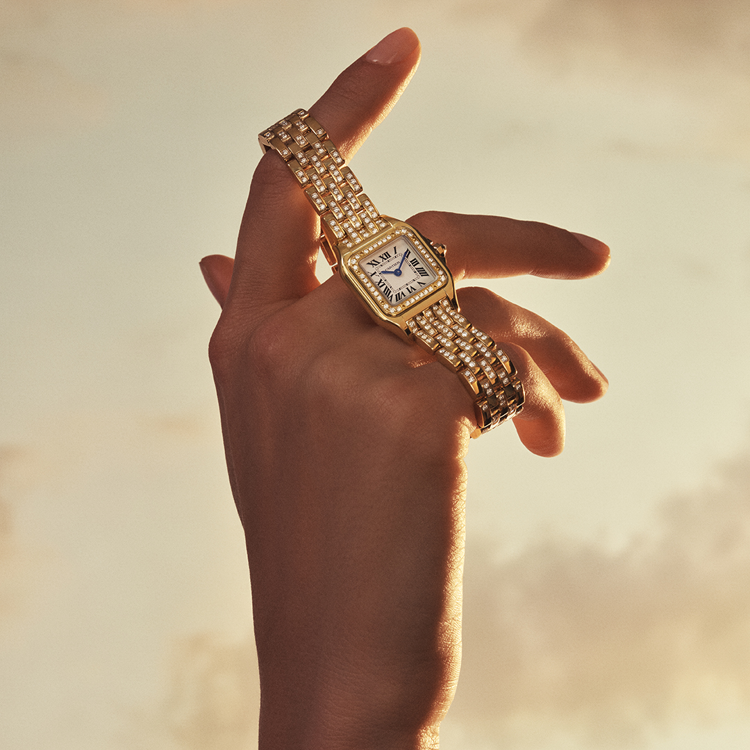 A hand wearing a gold and diamond-encrusted square watch against a cloudy sunset background.