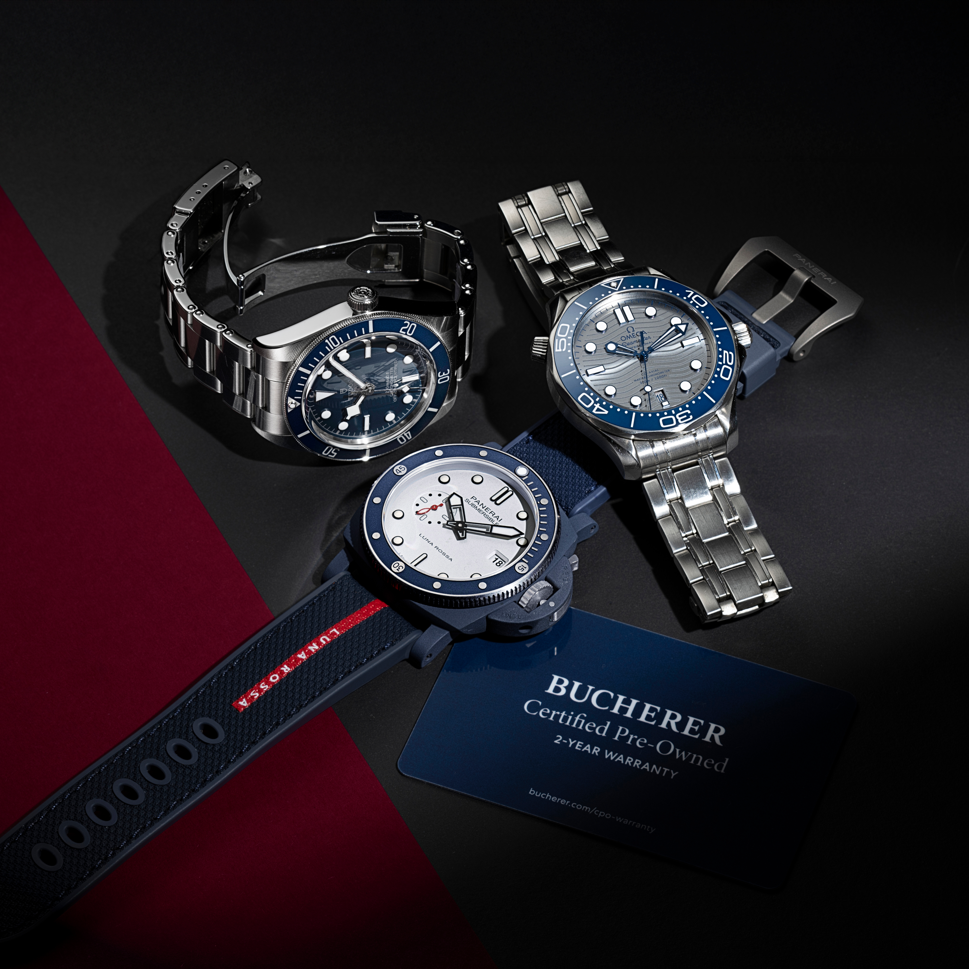 Trois montres-bracelets de luxe — deux avec bracelets en acier et une avec bracelet en caoutchouc bleu — sont exposées à côté d'une carte de garantie Bucherer.