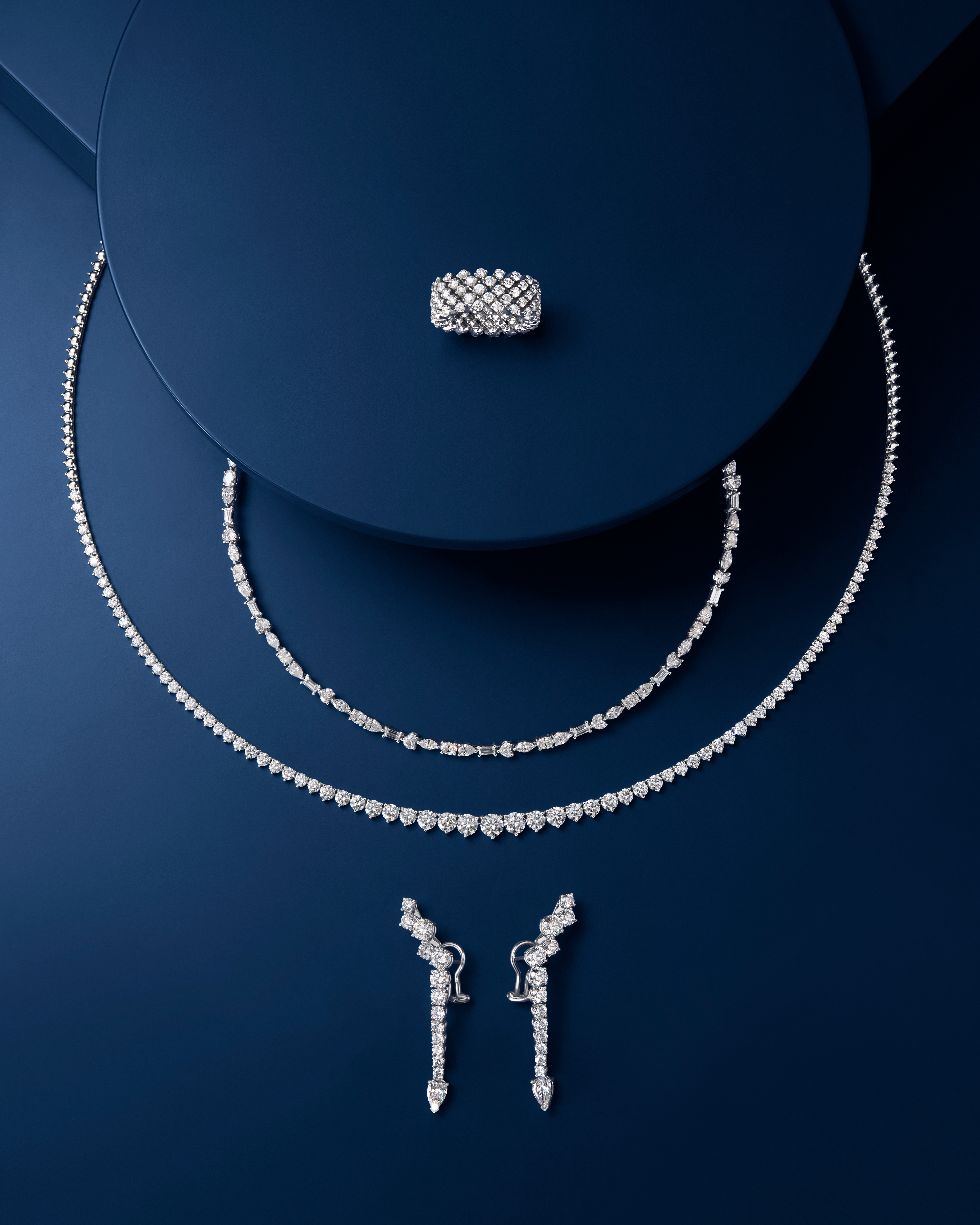 Ein Diamantschmuckset bestehend aus einem breiten Ring, zwei Halsketten und baumelnden Ohrringen, präsentiert auf dunkelblauem Hintergrund.
