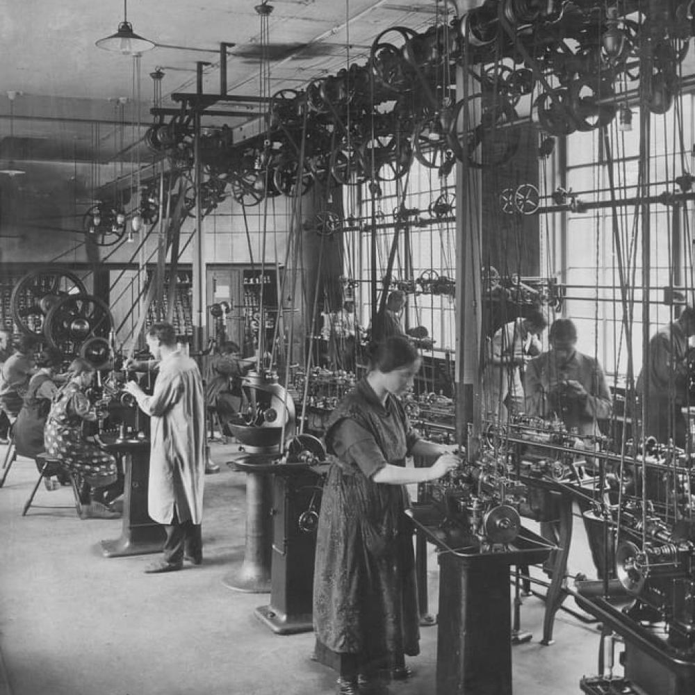 Black-and-white historical photo of workers operating machinery in a crowded textile factory powered by overhead belts.