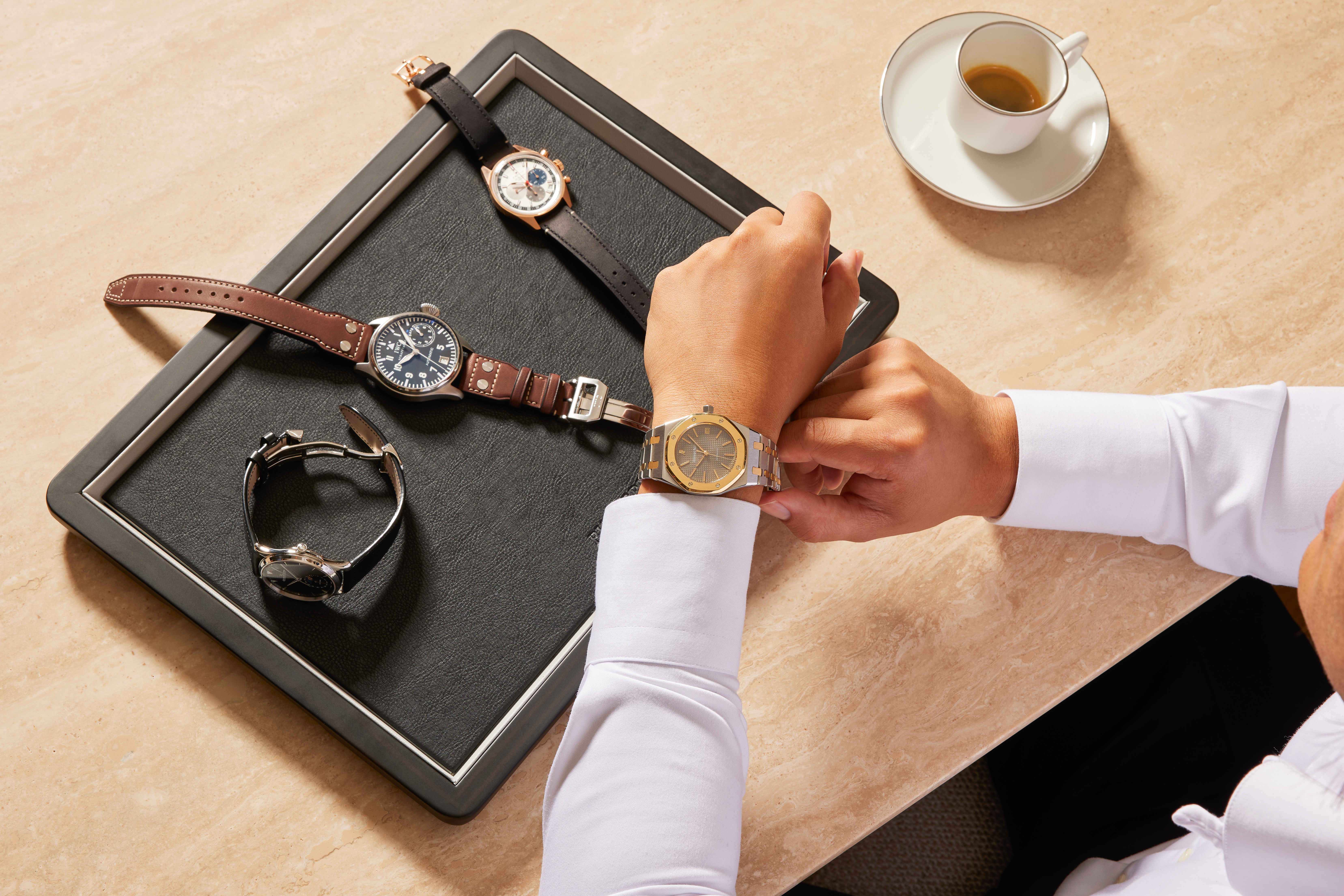 Eine Person probiert eine Uhr aus Gold und Stahl an; daneben liegen drei weitere Luxus-Armbanduhren auf einem schwarzen Tablett.