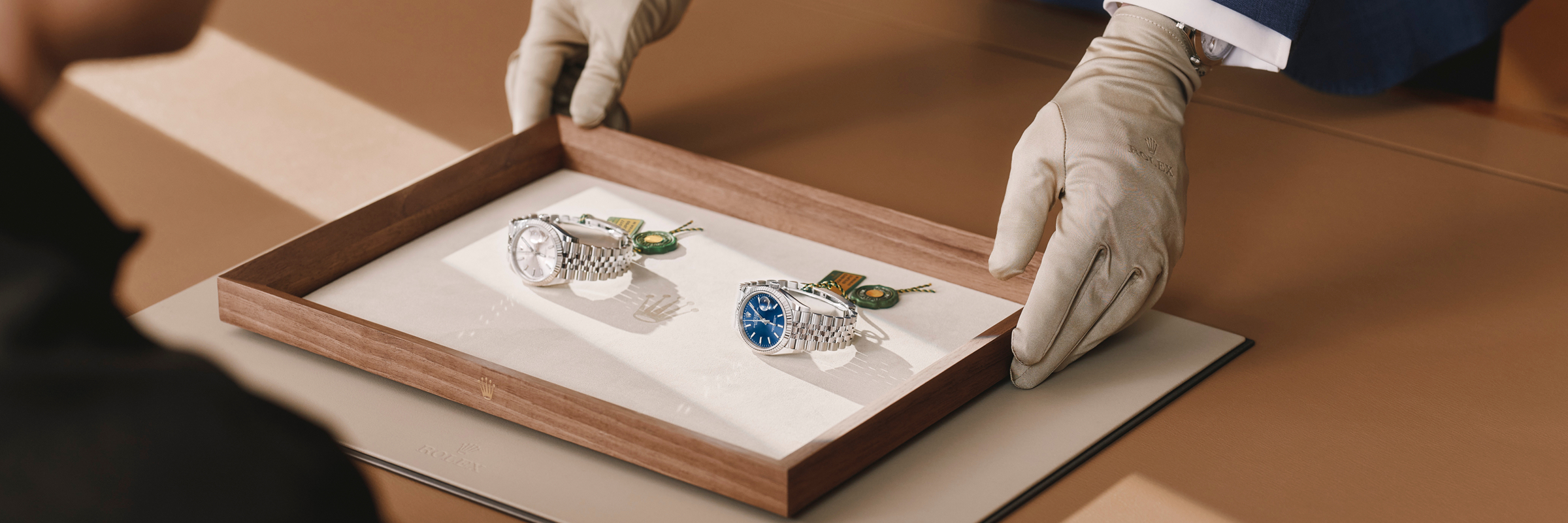 Eine Person mit weißen Handschuhen überreicht einem Kunden ein Holztablett mit zwei luxuriösen Rolex-Uhren.