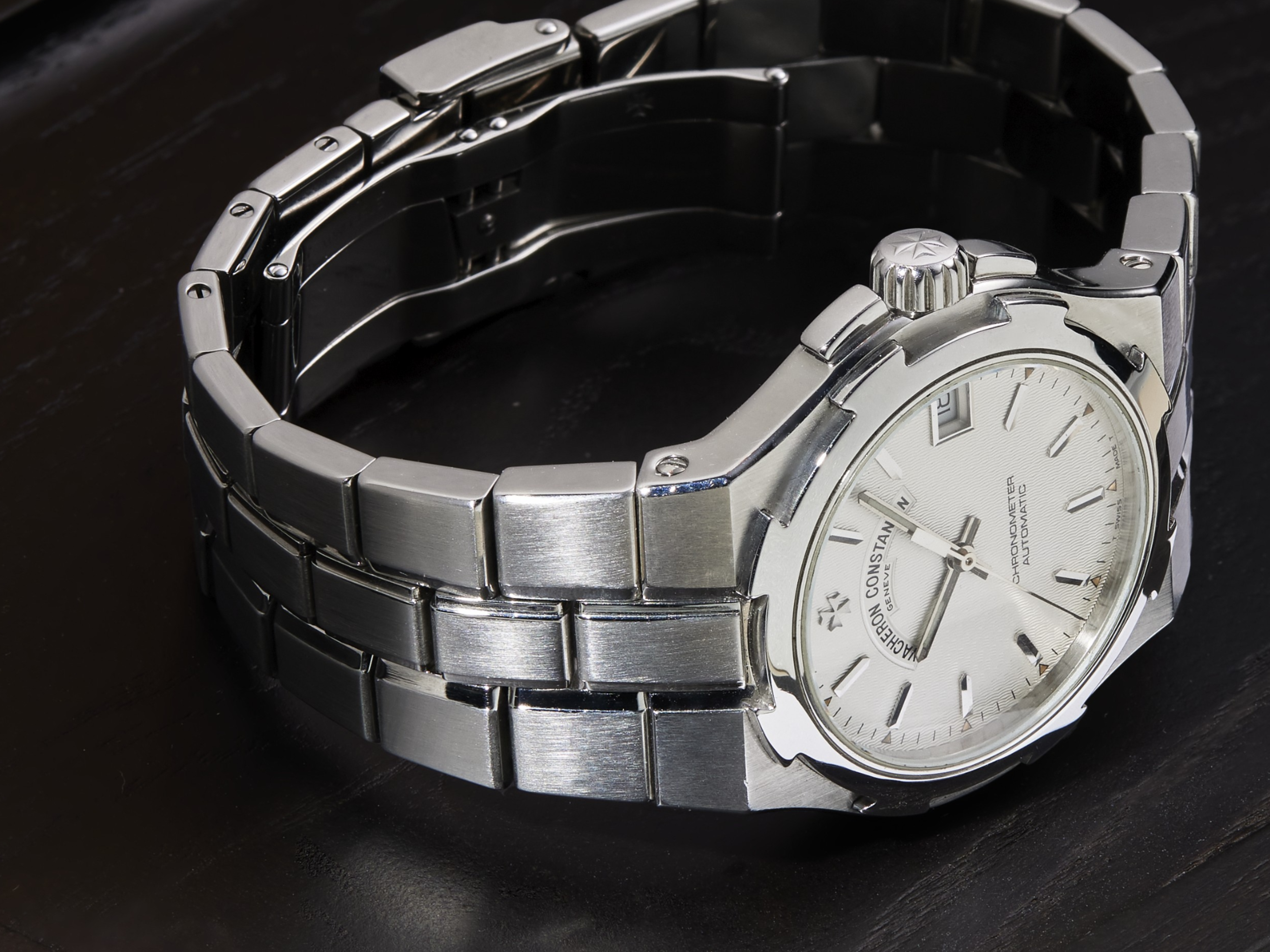 Eine Vacheron Constantin Armbanduhr aus Edelstahl mit weißem Zifferblatt und Gliederarmband aus Metall liegt auf einer dunklen Oberfläche.